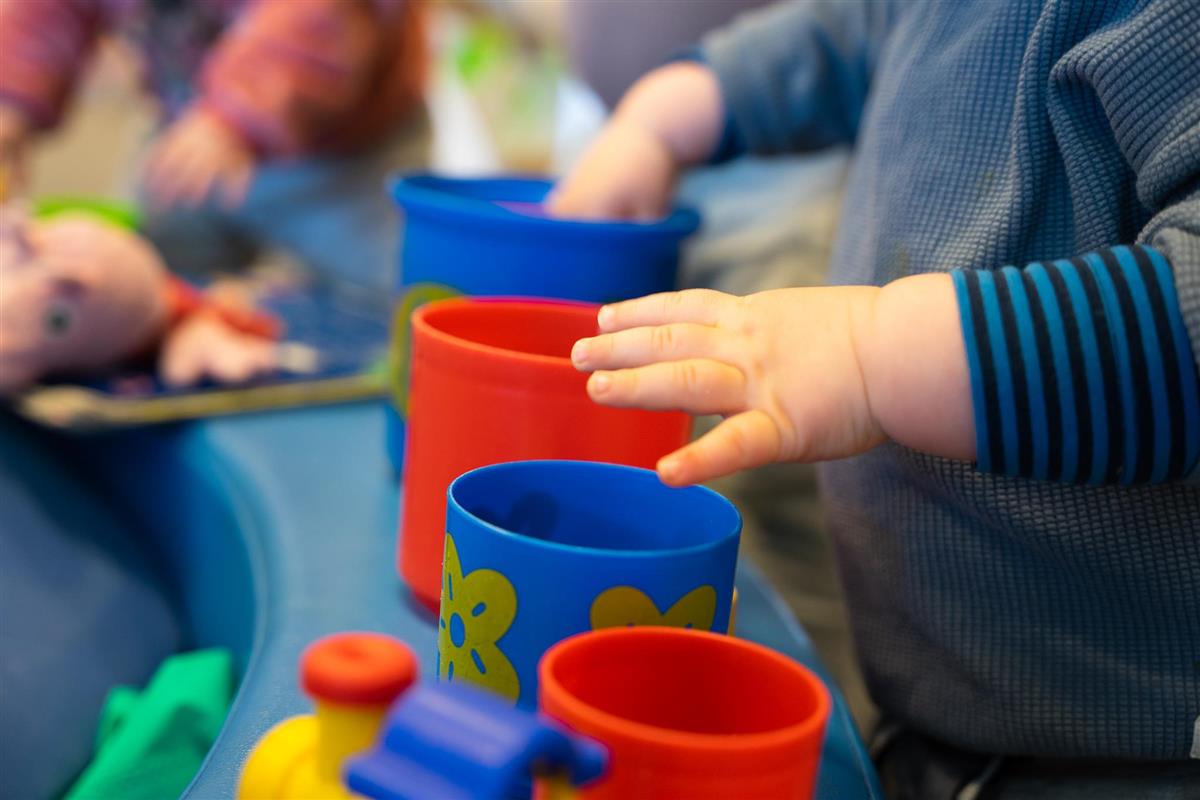 Barnehånd stabler fargerike plastkopper på en blå overflate. Rosa kosedyr er synlig i nærheten. Uskarp bakgrunn - Klikk for stort bilete